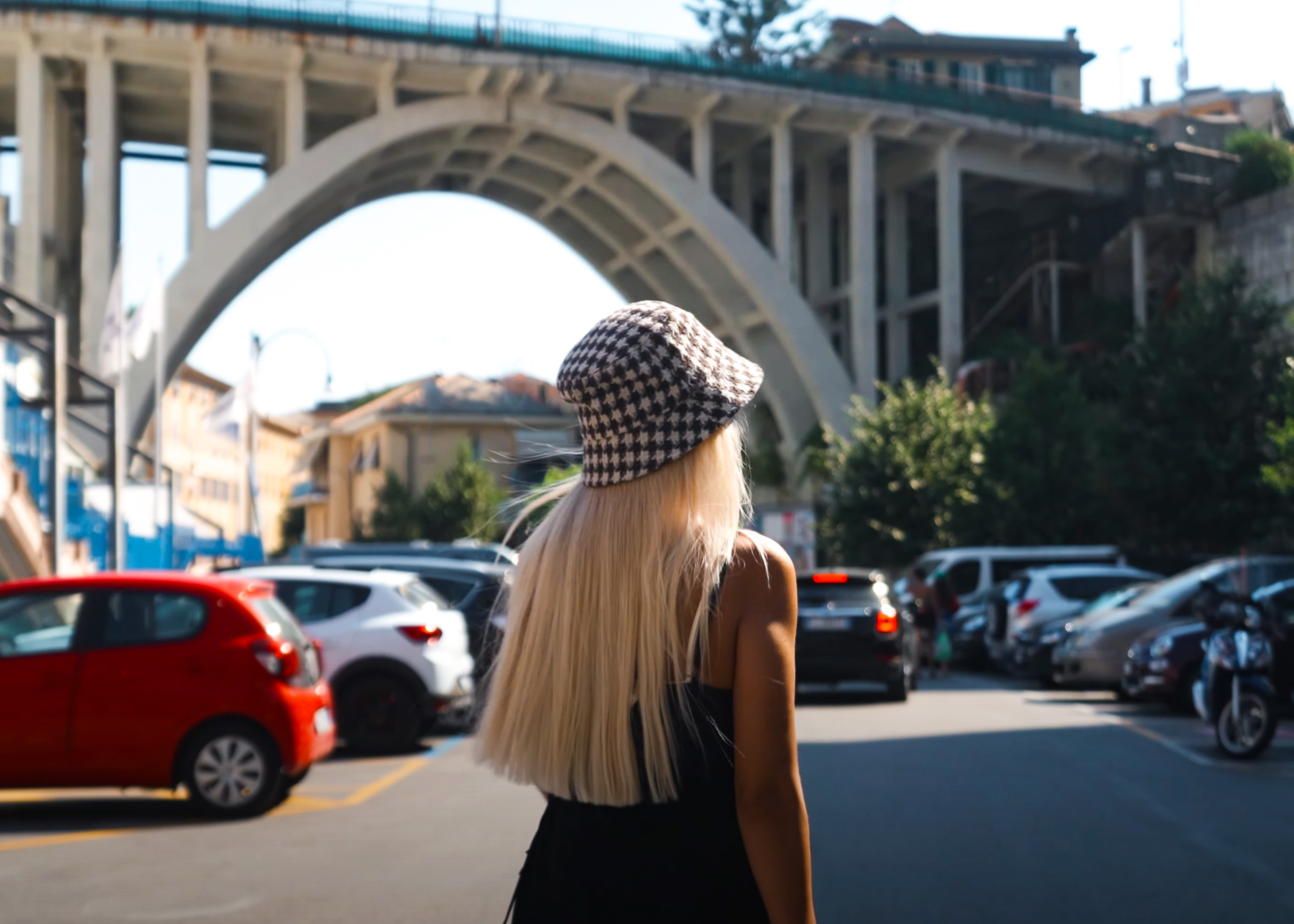 Woman with long blonde hair wearing a checkered bucket hat, walking toward the large arched bridge in Bogliasco, Italy.