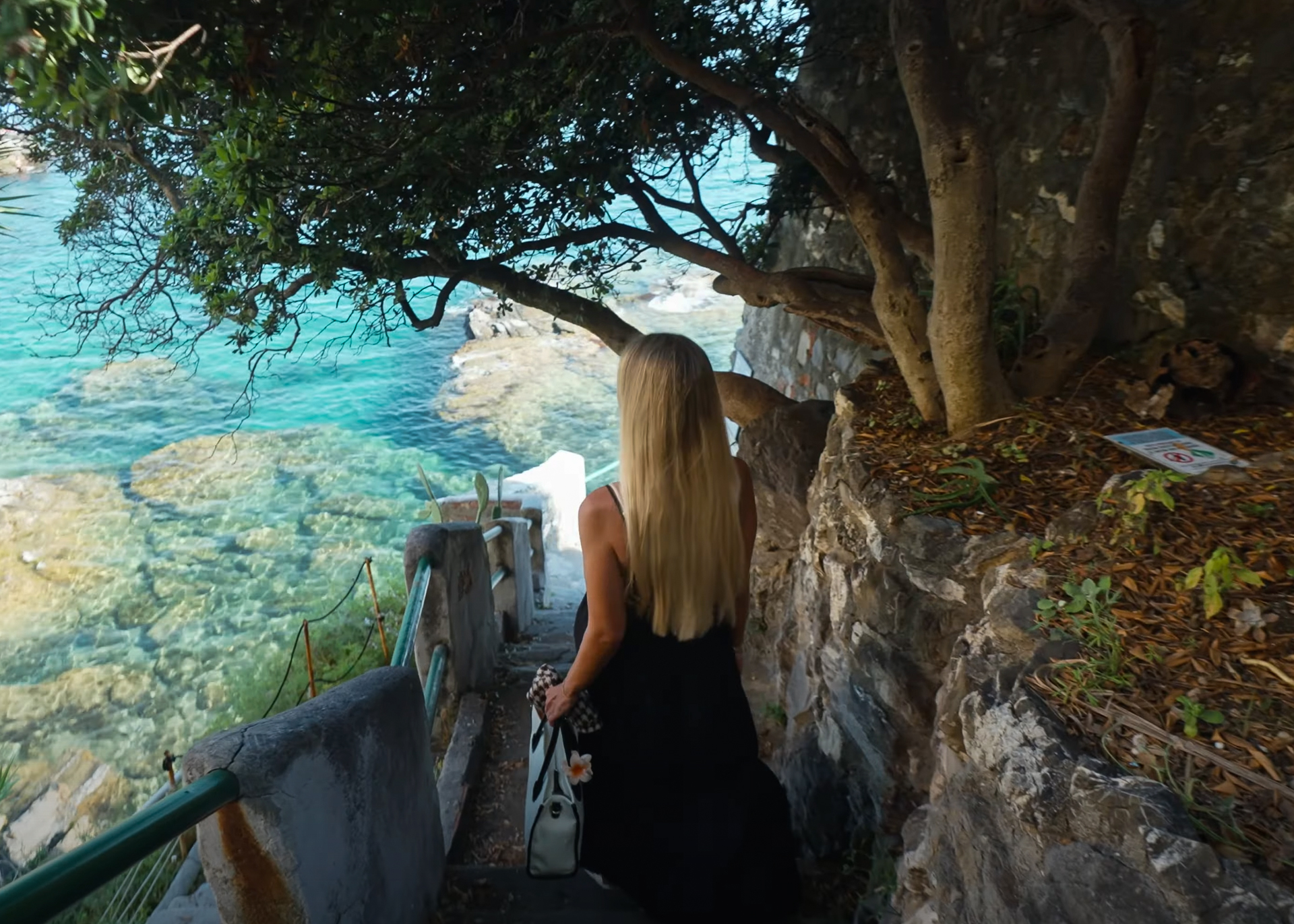 Woman with long blonde hair walking down stone steps shaded by trees toward Scogliera di Pontetto the turquoise sea in Bogliasco, Italy.