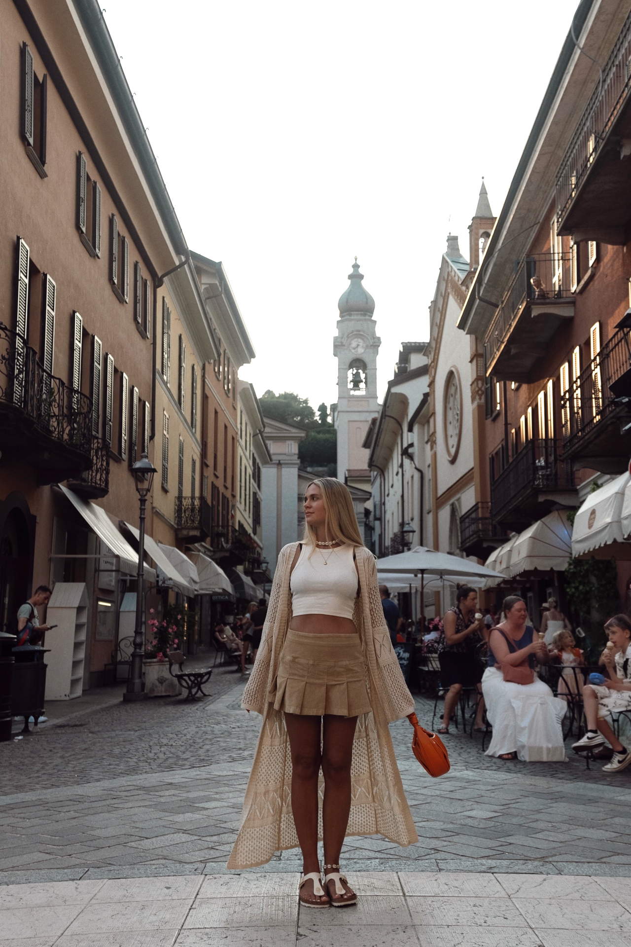 Stylish woman standing in the old town of Menaggio with church tower and lively cafés in the background, Lake Como, Italy.