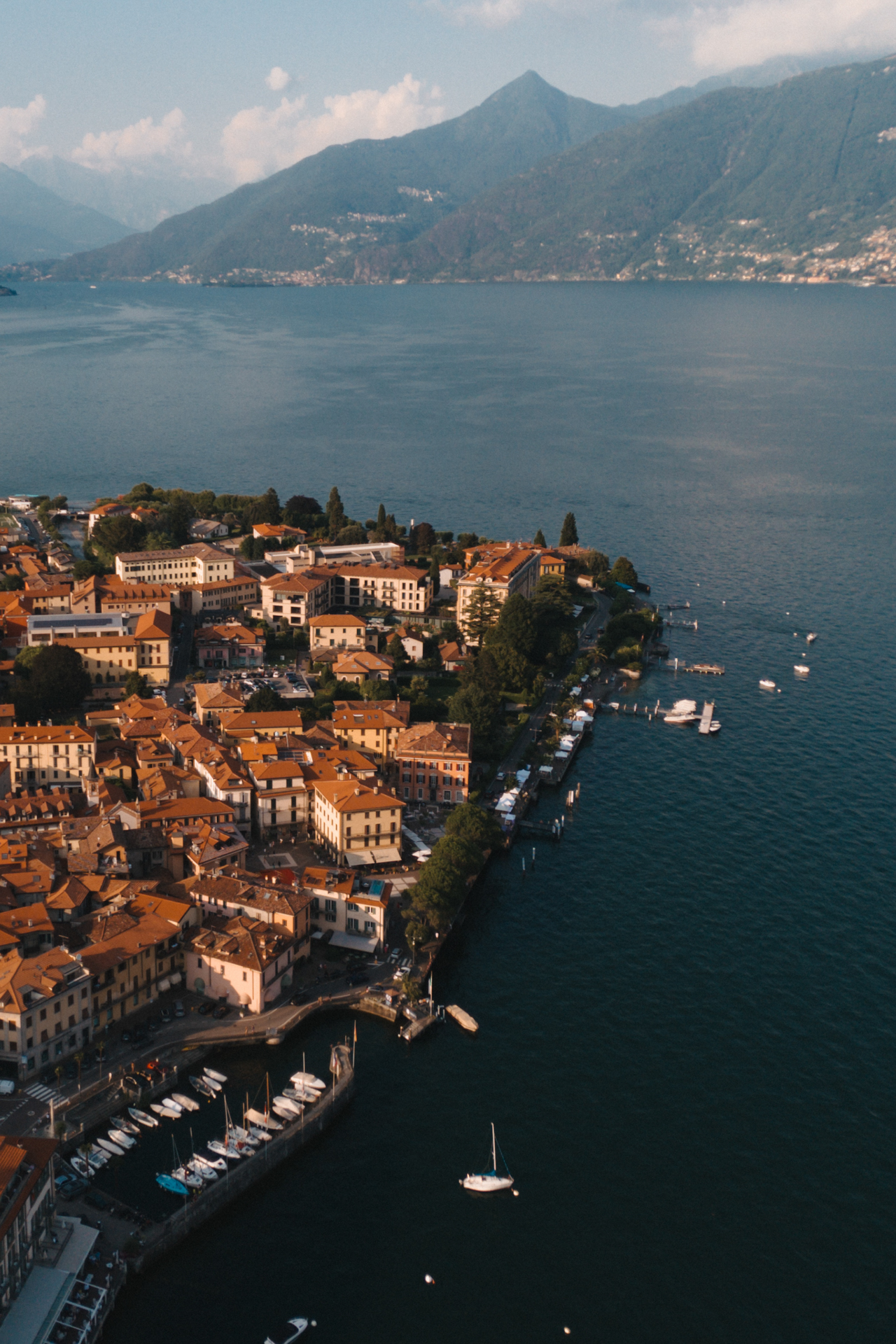 Aerial drone view of Menaggio town and Lake Como shoreline with colorful houses, marina, and surrounding mountains.