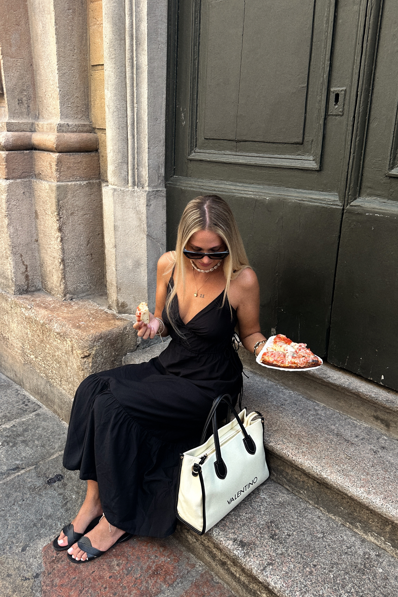 Woman in black dress sitting on steps in Milan eating pizza with Valentino bag