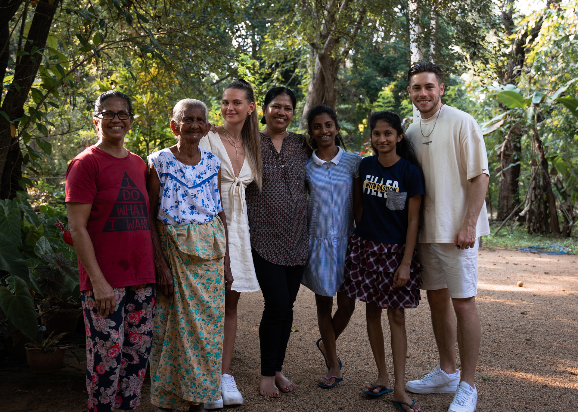 A group photo of a smiling Sri Lankan family with travelers, standing together outdoors in a lush garden.