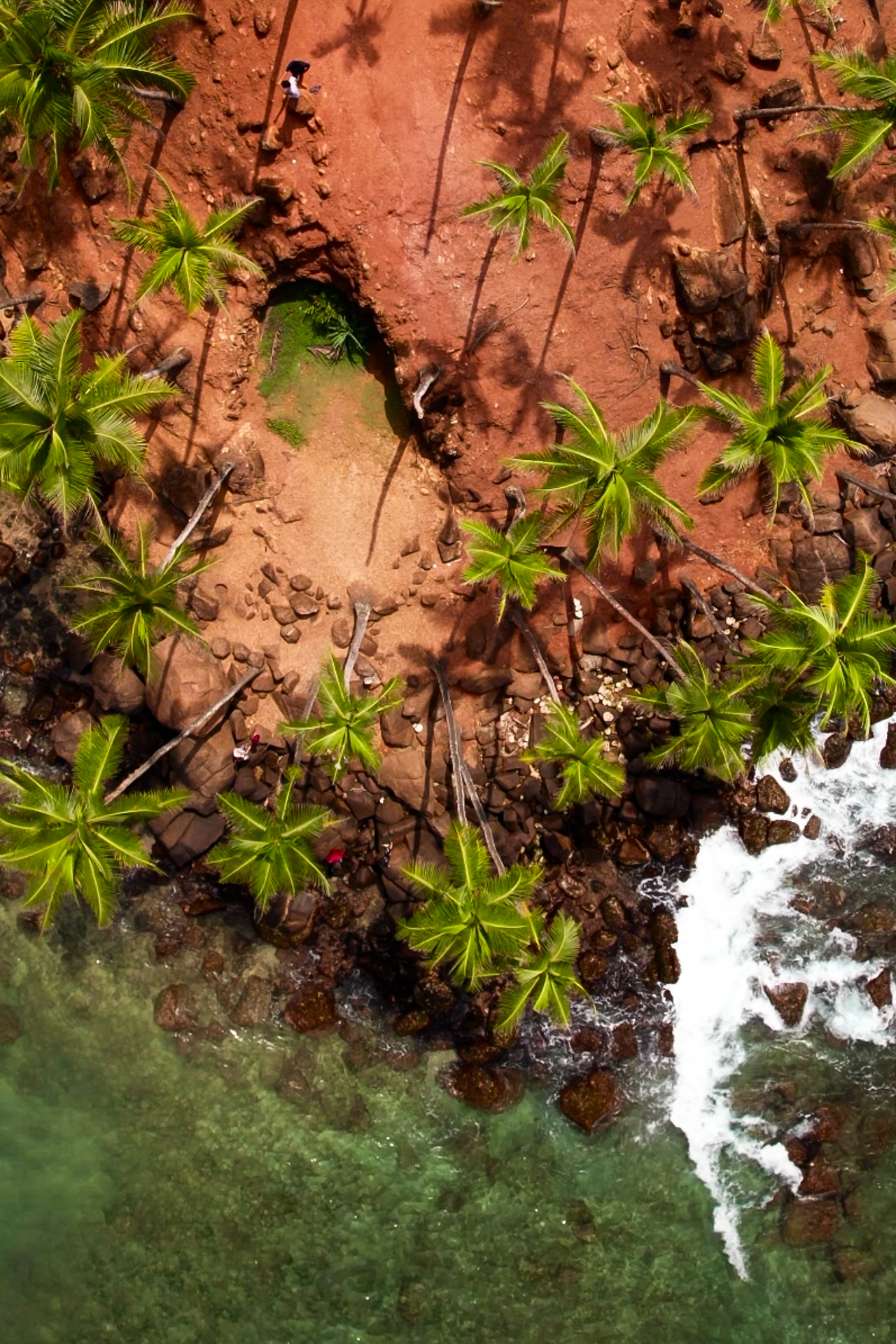 Drone view of Coconut Tree Hill in Mirissa, showing red soil, palm trees, and turquoise waves crashing below.