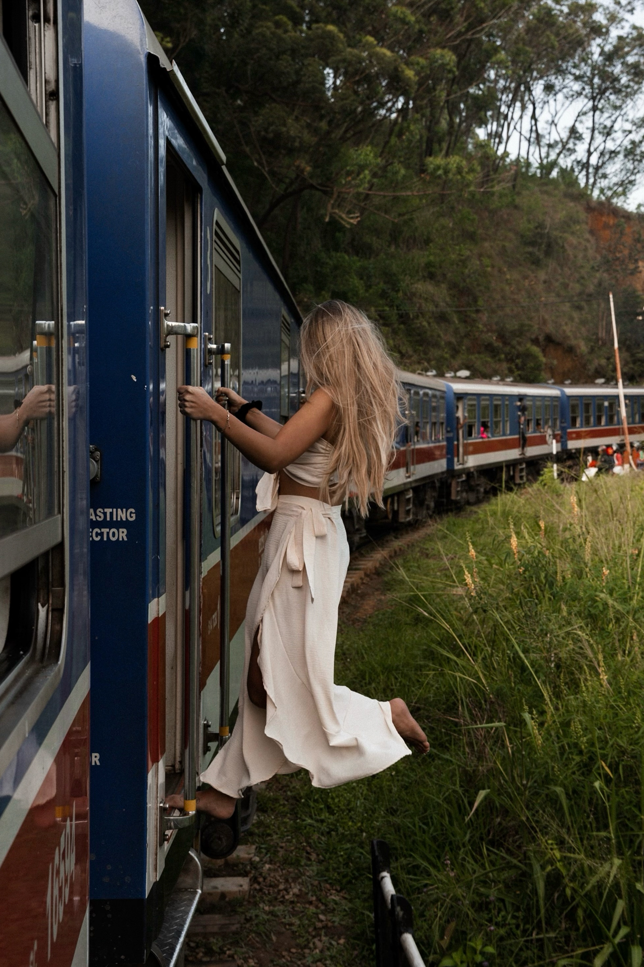 A woman holding onto the doorway of the famous blue train in Sri Lanka as it winds through the lush highlands.