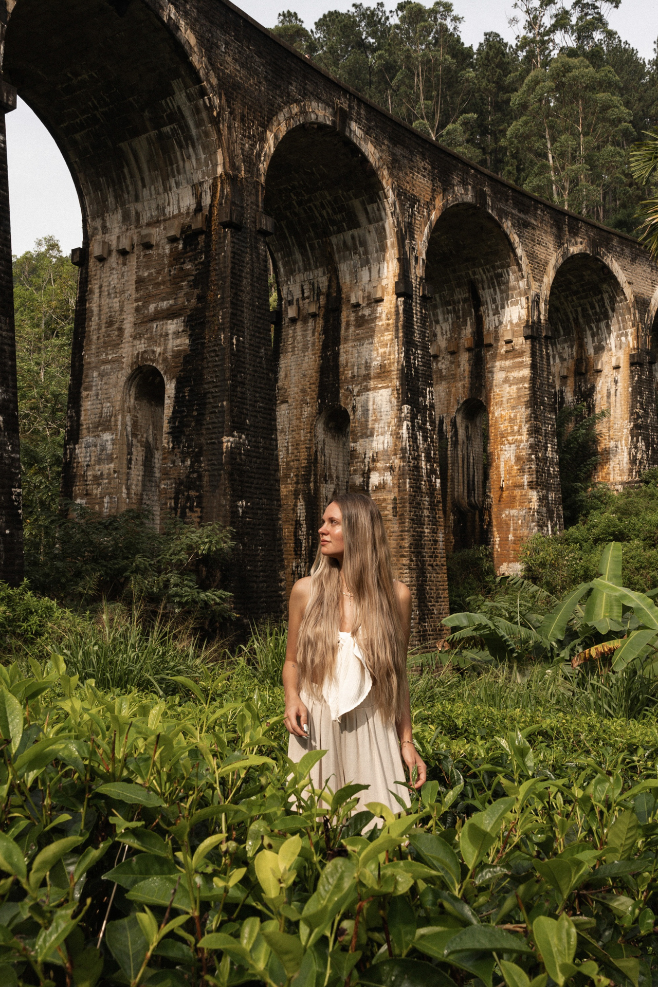 Woman standing in front of the iconic Nine Arches Bridge surrounded by lush greenery in Ella, Sri Lanka.