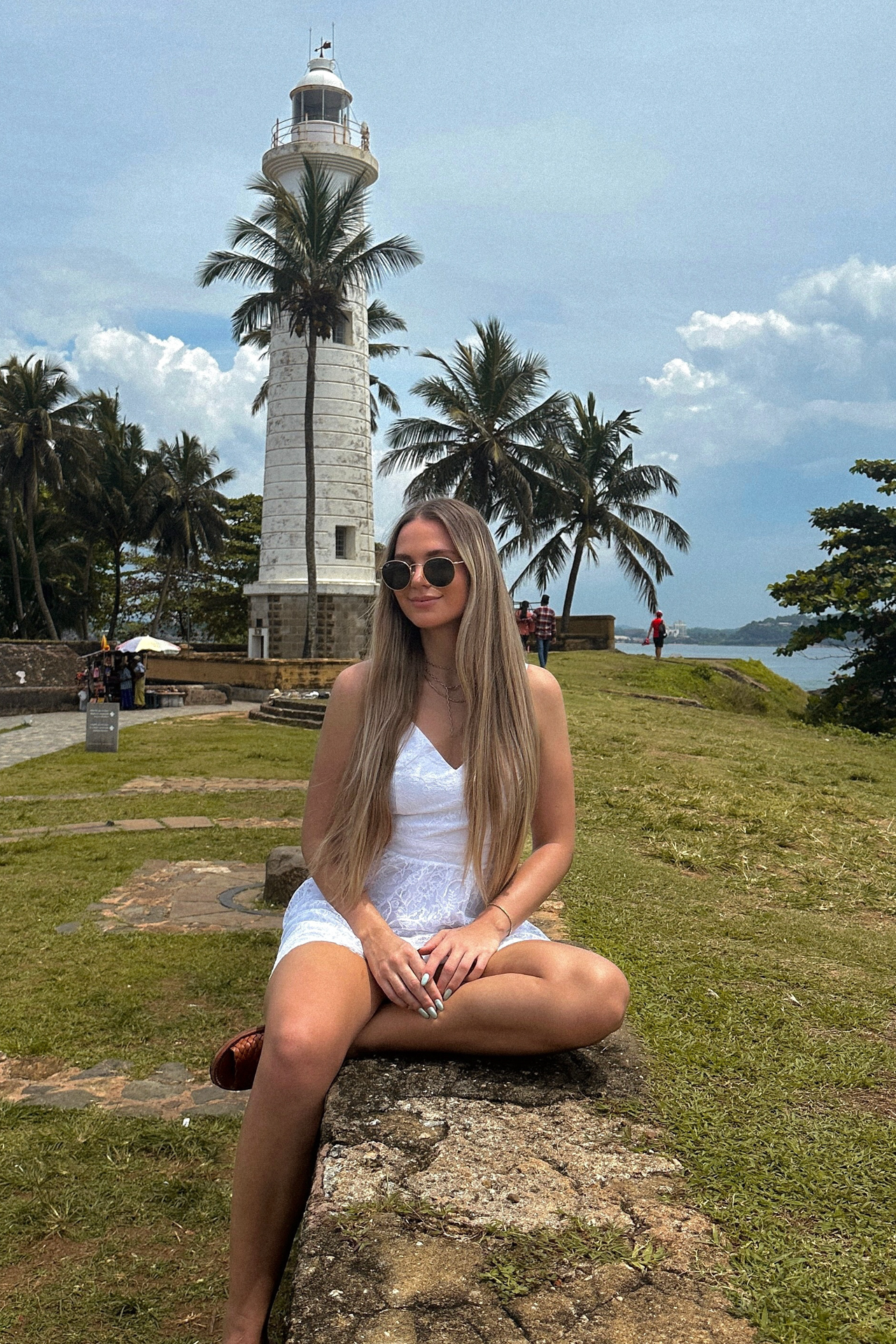 Woman sitting on a stone wall with the Galle Lighthouse and palm trees rising behind her.