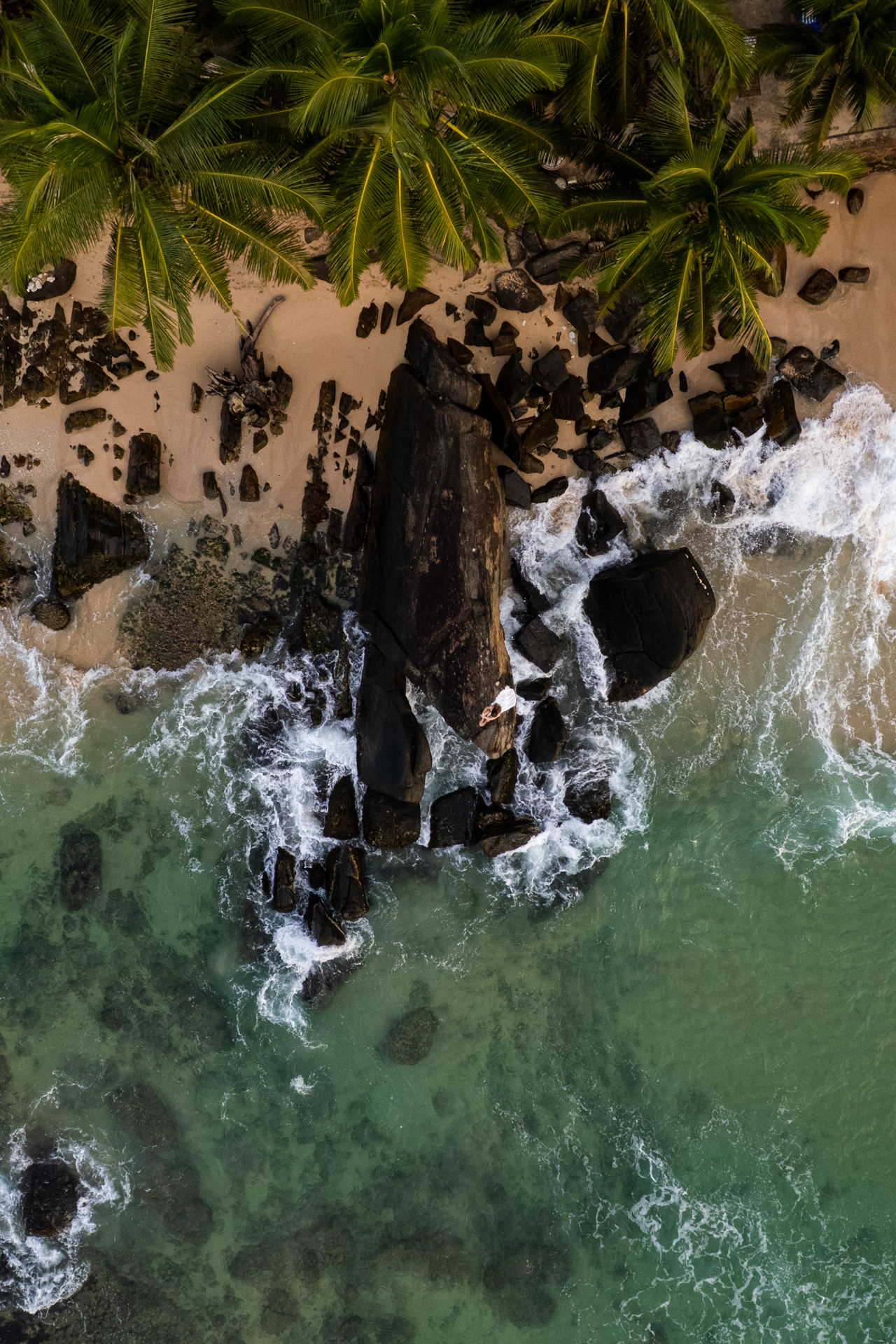 Aerial view of a rocky coastline in Sri Lanka with turquoise waves crashing against black rocks and palm trees lining Dalawella beach.