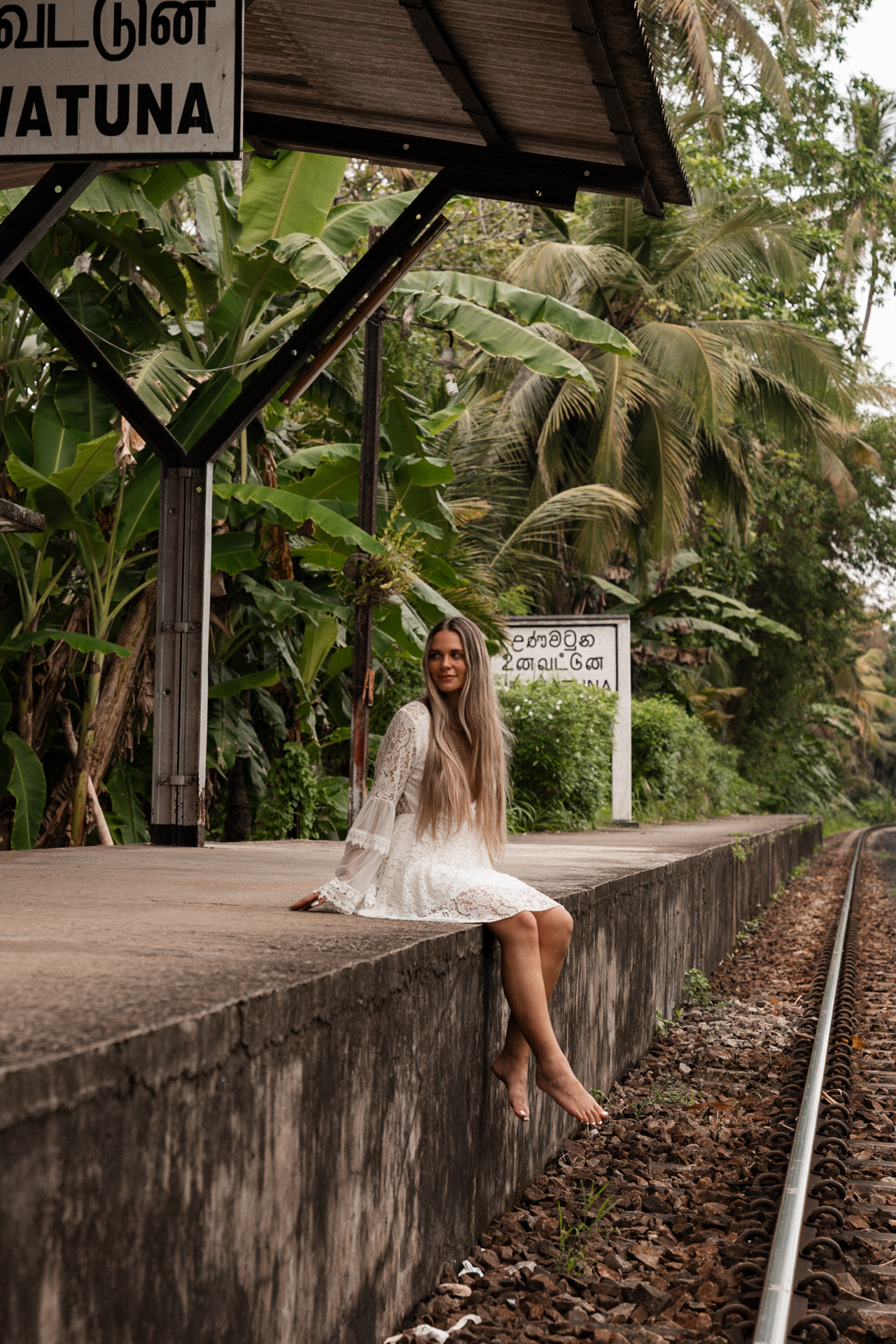 Woman sitting barefoot on the platform at Unawatuna train station in Sri Lanka, surrounded by tropical greenery.