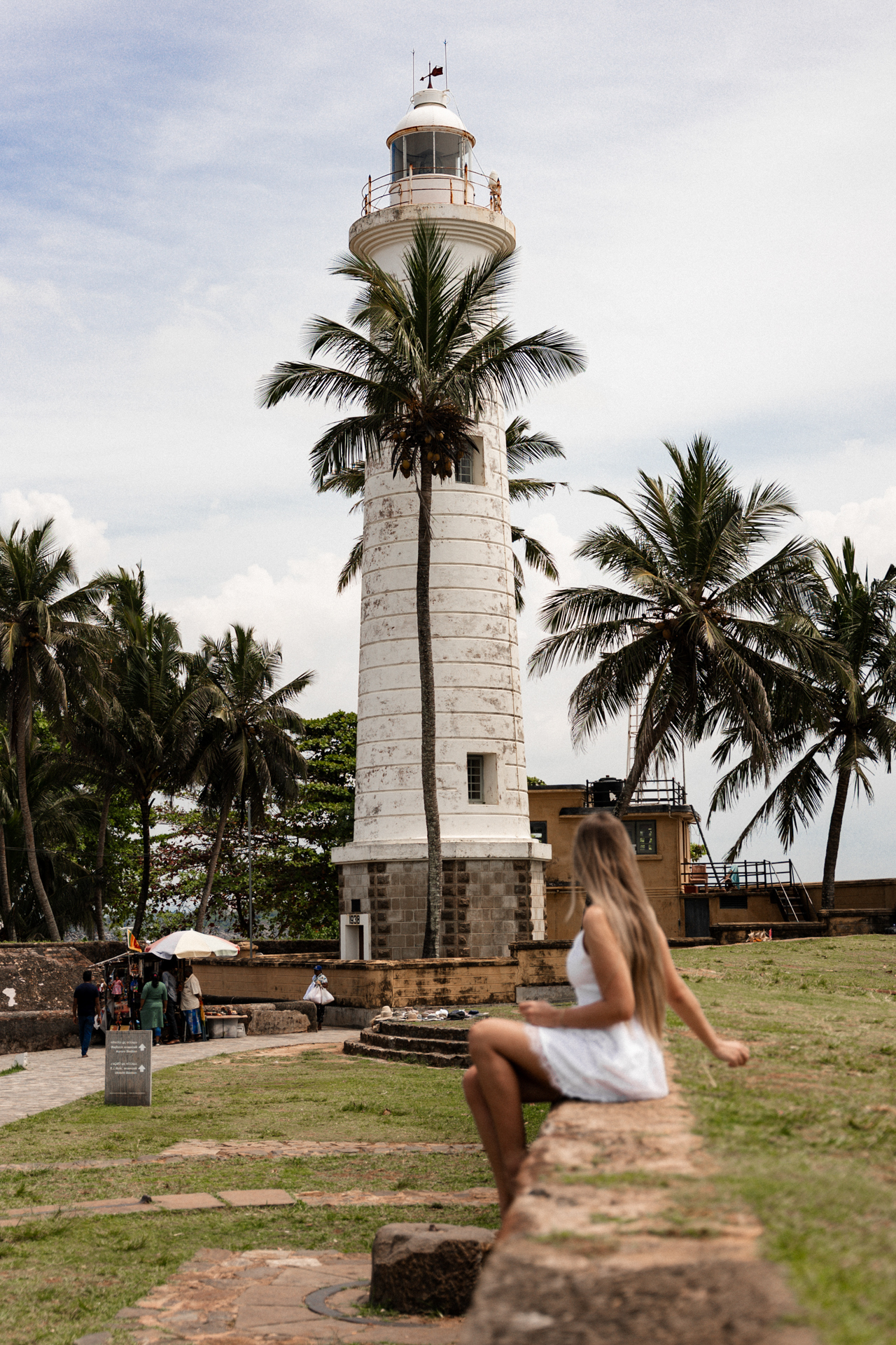 Woman sitting on a stone wall with the Galle Lighthouse and palm trees rising behind her.