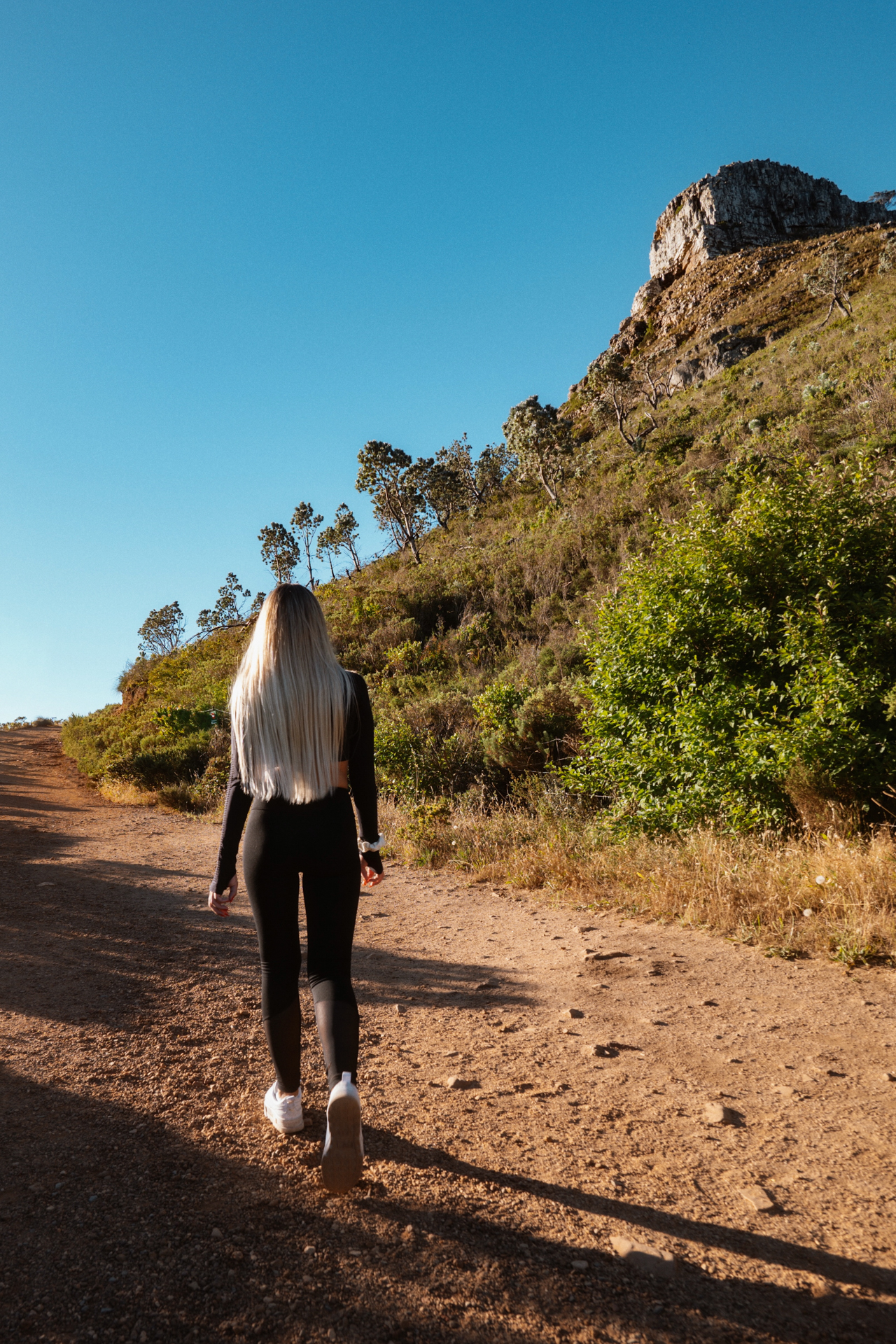 Lion's Head Woman hiking uphill trail with rocky cliffs and blue sky in Cape Town.