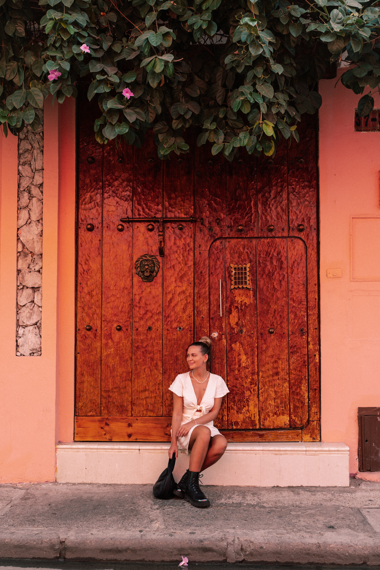 Cartagena Woman sitting in front of a large wooden colonial door framed by pink walls and flowers in Cartagena, Colombia.