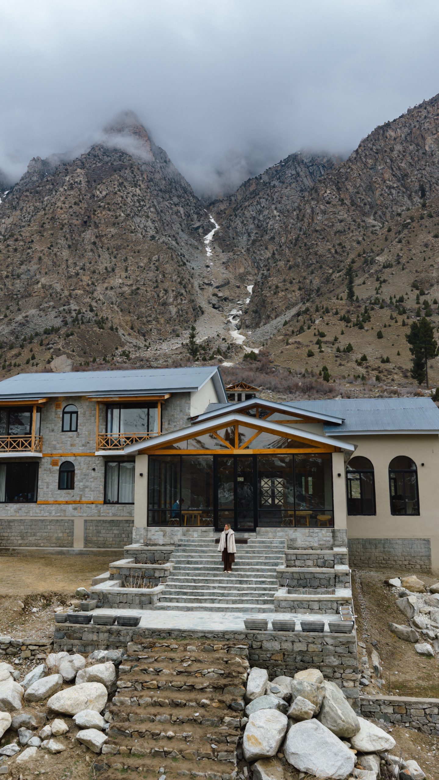 Naltar Valley Mountain lodge in Gilgit-Baltistan, Pakistan, with a woman standing on stone steps and misty peaks in the background.