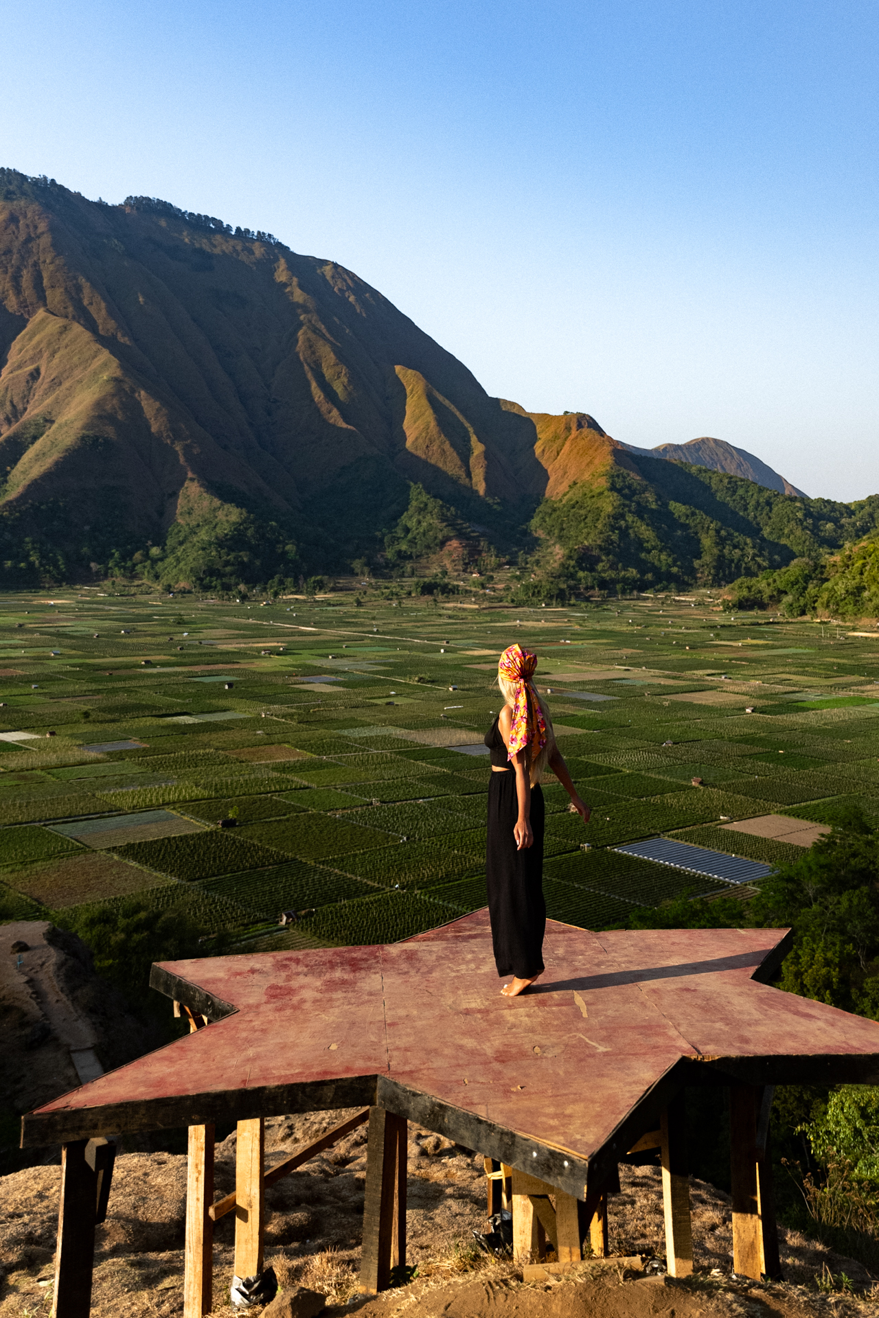 Lombok Woman standing on a star-shaped wooden platform overlooking vast green rice fields and Lombok’s dramatic mountains.