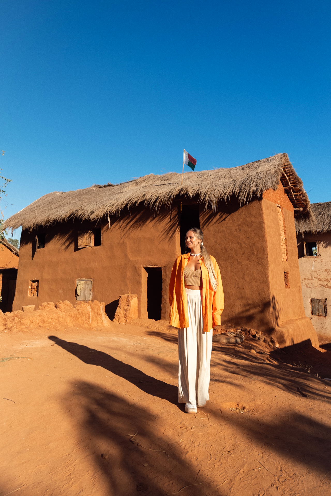 Madagascar Road Trip Traveler standing in front of a traditional clay house with a thatched roof and Madagascar flag under clear blue sky.