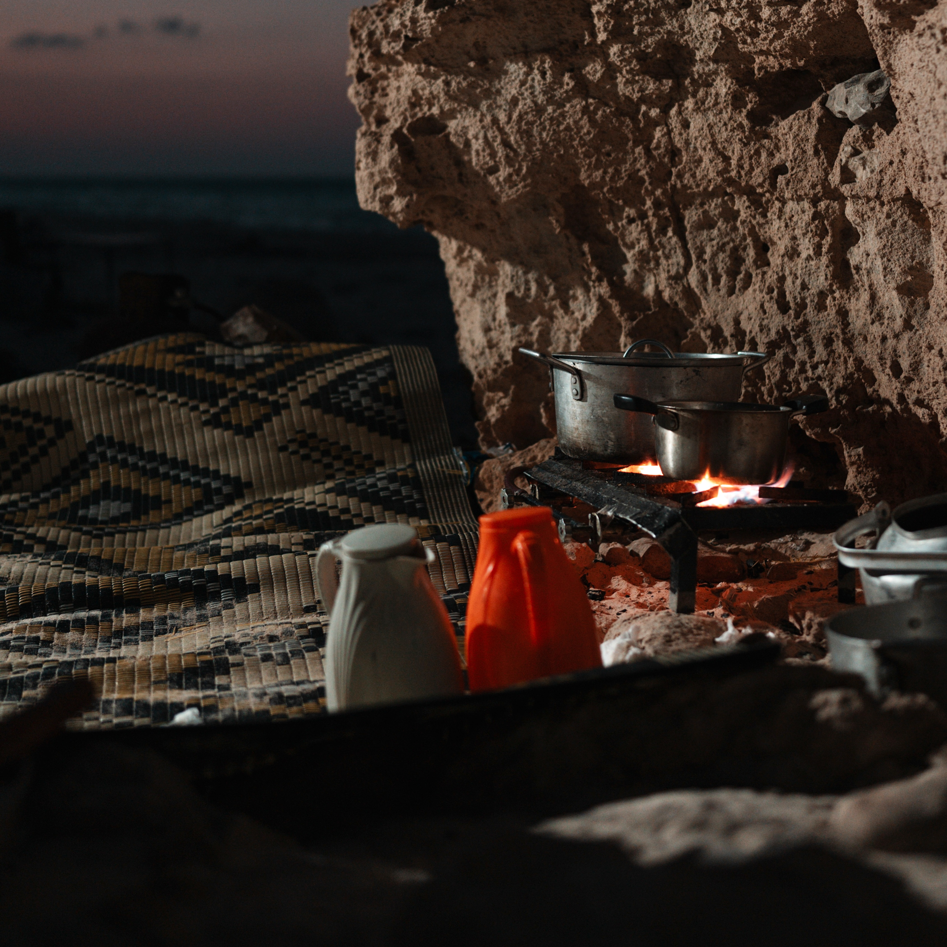 Food in Socotra Cooking pots over an open fire in a rocky cave on Socotra, with a mat and jugs nearby at dusk.