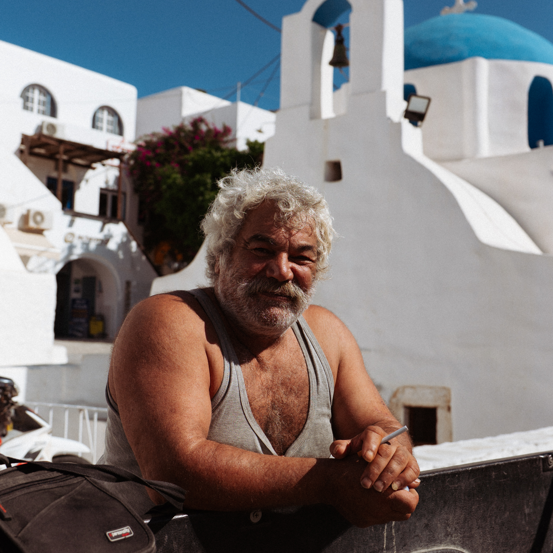 Culture in Greece A Greek man with curly white hair and a mustache sits outside in Ios, with a traditional whitewashed church and blue dome in the background.
